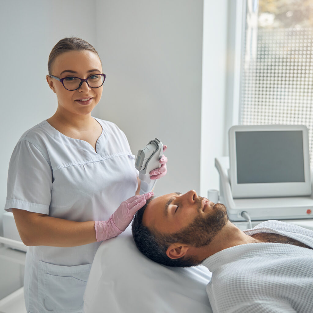 female esthetician holding a laser device and smiling while male patient lying on daybed