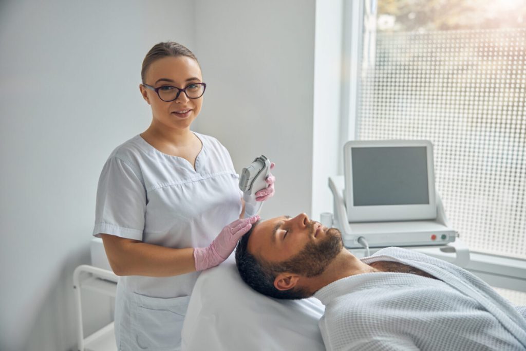 female esthetician holding a laser device and smiling while male patient lying on daybed